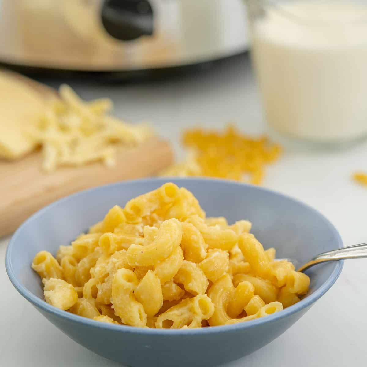 A two tone ceramic bowl of macaroni cheese sitting in front of ingredients and a slow cooker.