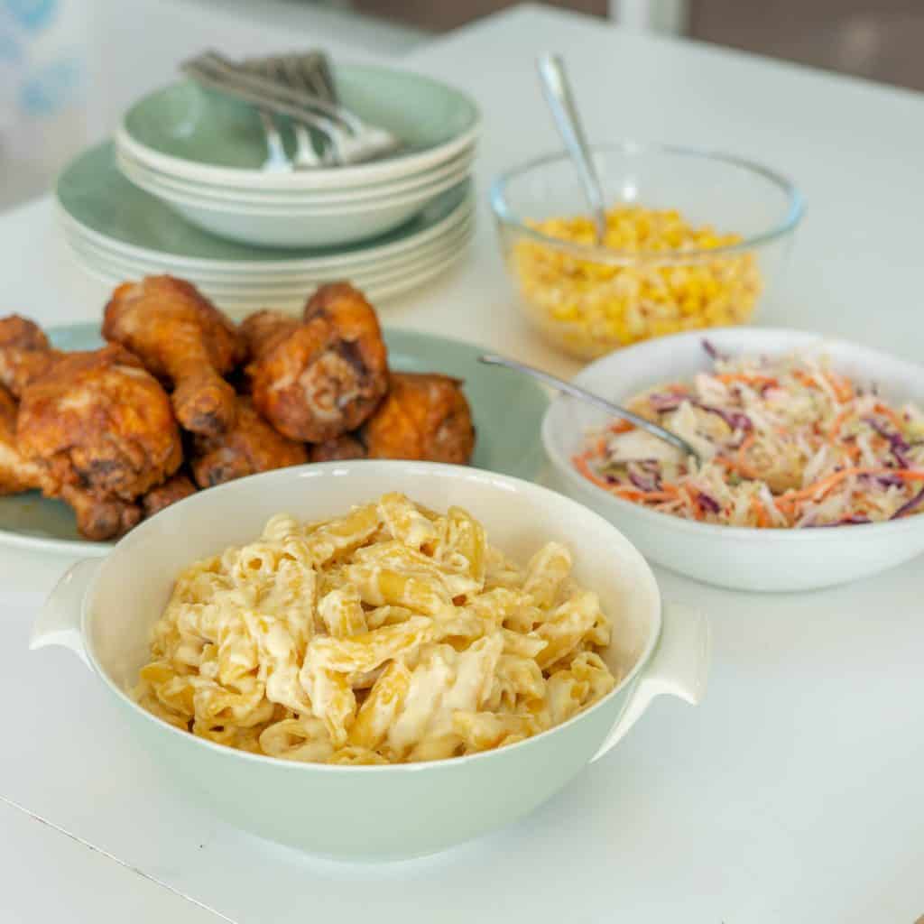 Cheese pasta, coleslaw, chicken drumsticks, and corn kernels on a table.