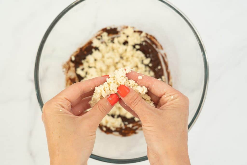Hands with orange nail polish crumbling firm tofu into a large bowl containing spices.