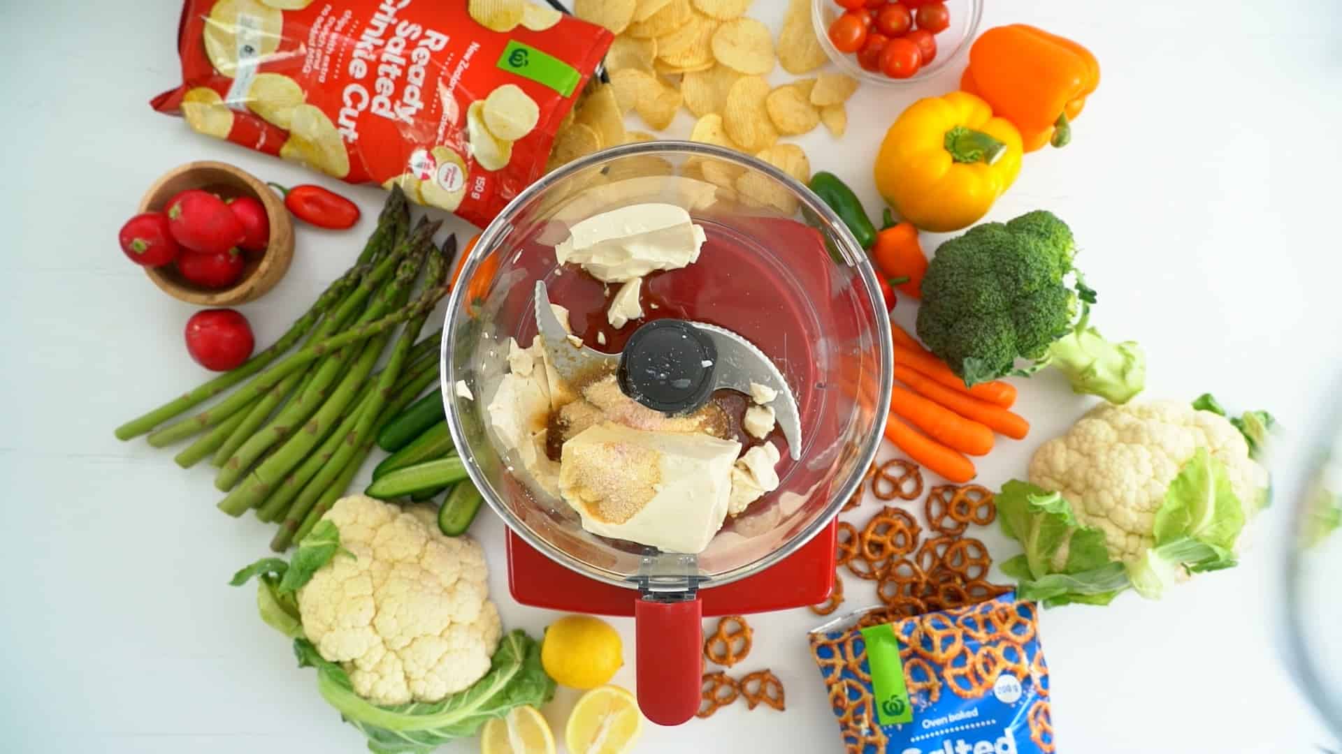 Tofu, onion powder and soy sauce in food processor sitting on a bench with fresh produce.
