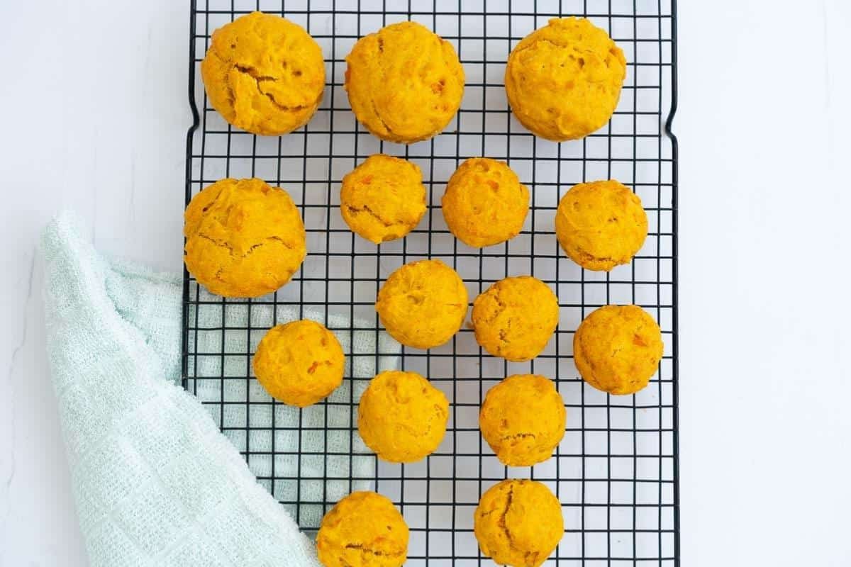 Mini carrot muffins and standard sized carrot muffins colling on a black wire rack with a mint tea towel draped over the corner of the rack.