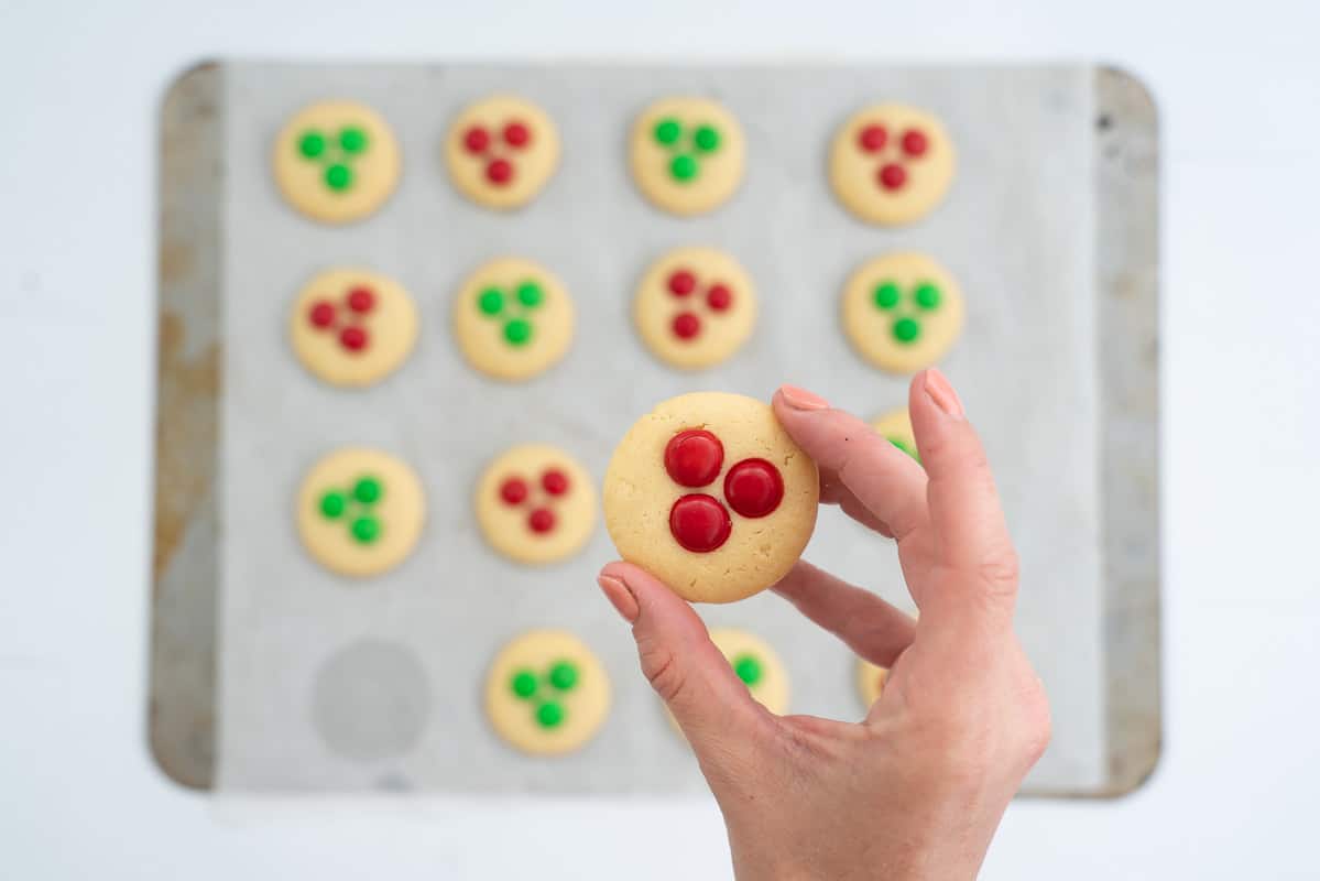 A hand holding a m and m decorated cookie above a cookie tray.