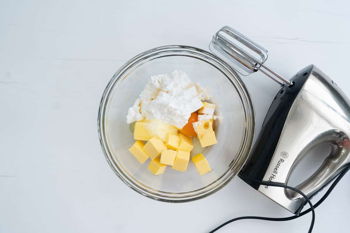 A glass mixing bowl with butter, vanilla essence, eggs and icing sugar in it, next to a hand held electric beater.