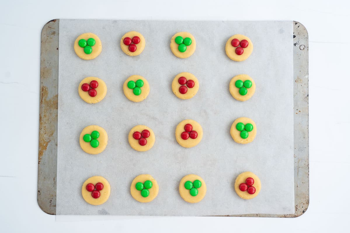 Cookies ready to go into the oven on a lined cookie tray, each cookie is topped with 3 red or green m and ms.