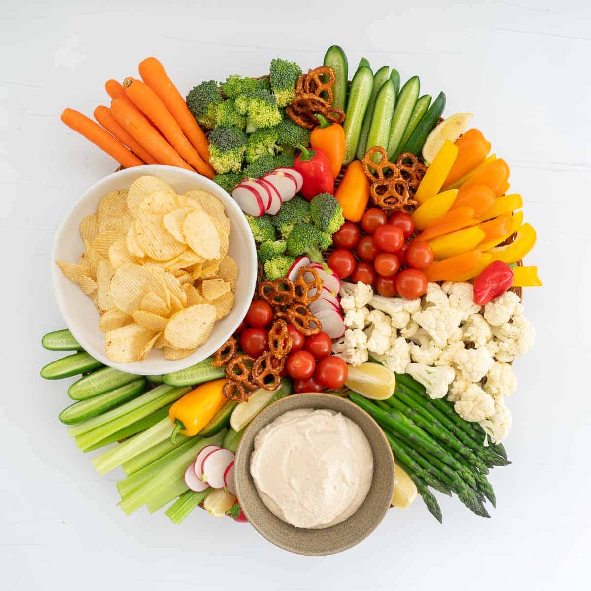 A large round platter of vegetable crudité, potato crisps, pretzels and onion dip. ,