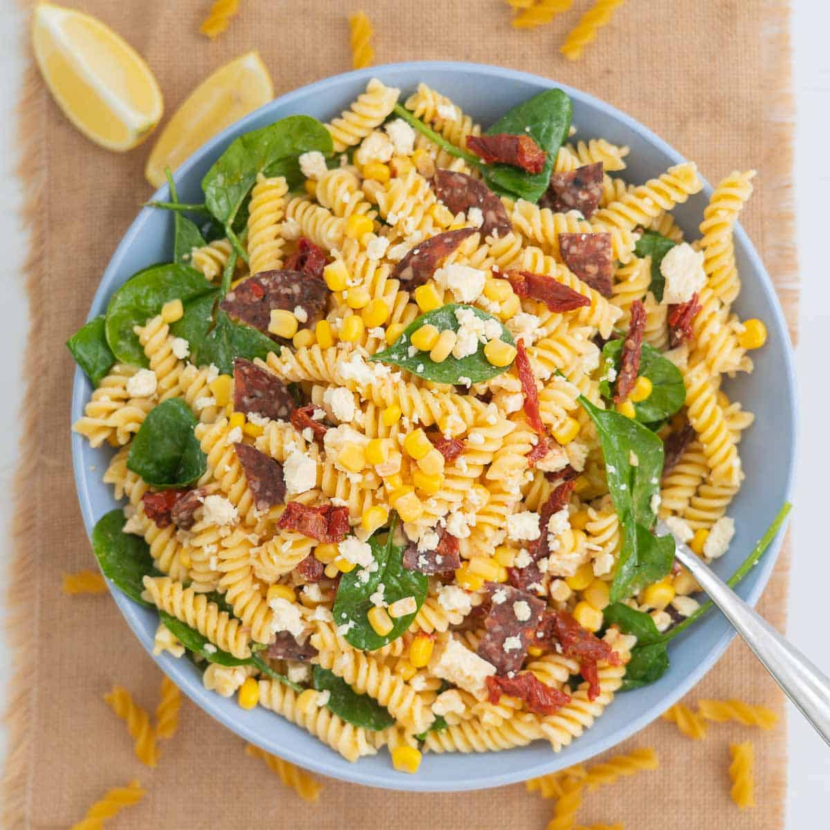 A large bowl filled with pasta, salami, sundried tomatoes, feta and spinach leaves, sitting on a hesian table runner scattered with lemon wedges.
