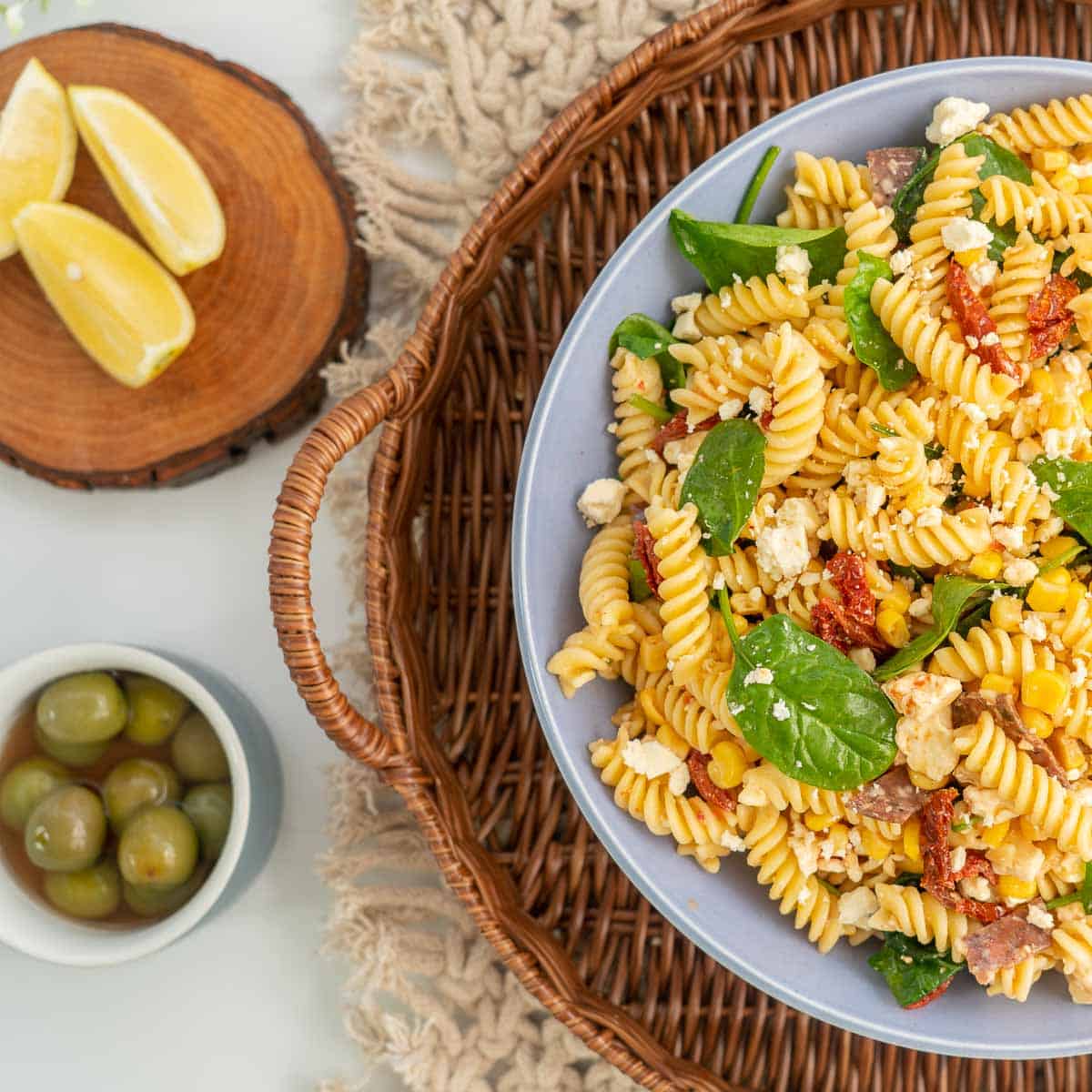 A large bowl of pasta salad with smaller bowls of green olives, lemon wedges and pinenuts.