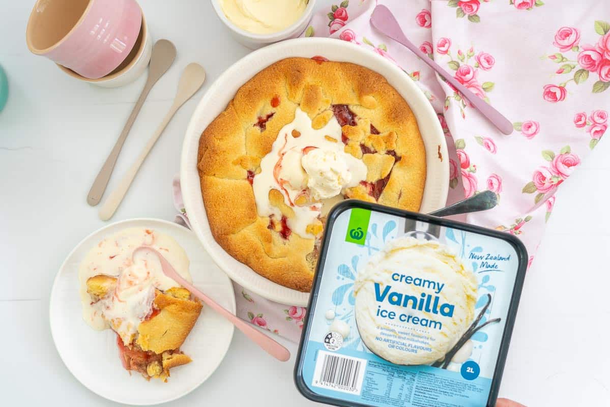 A rhubarb pie in a white pie dish, topped with a scoop of melted ice cream. The lid for the creamy vanilla ice cream is being held above the pie.