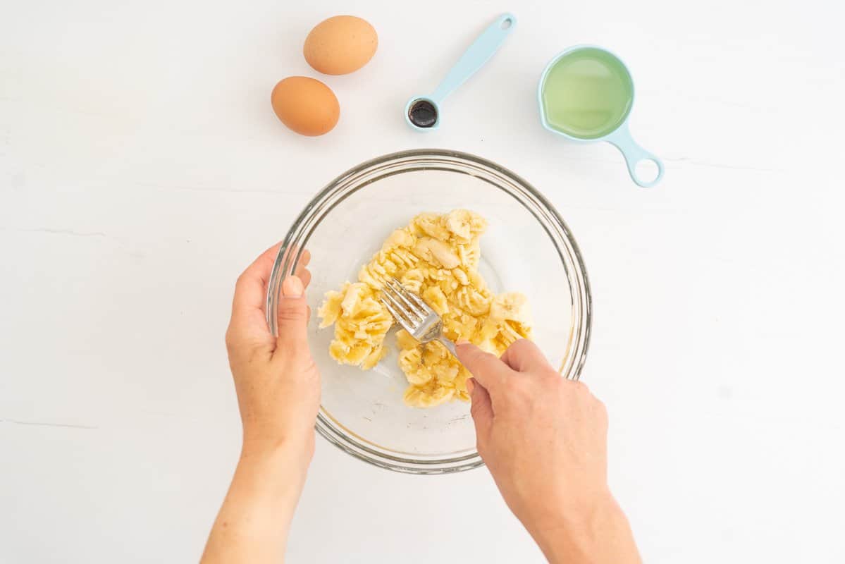 Bananas being mashed in a glass bowl, other muffin ingredients on the bench top.