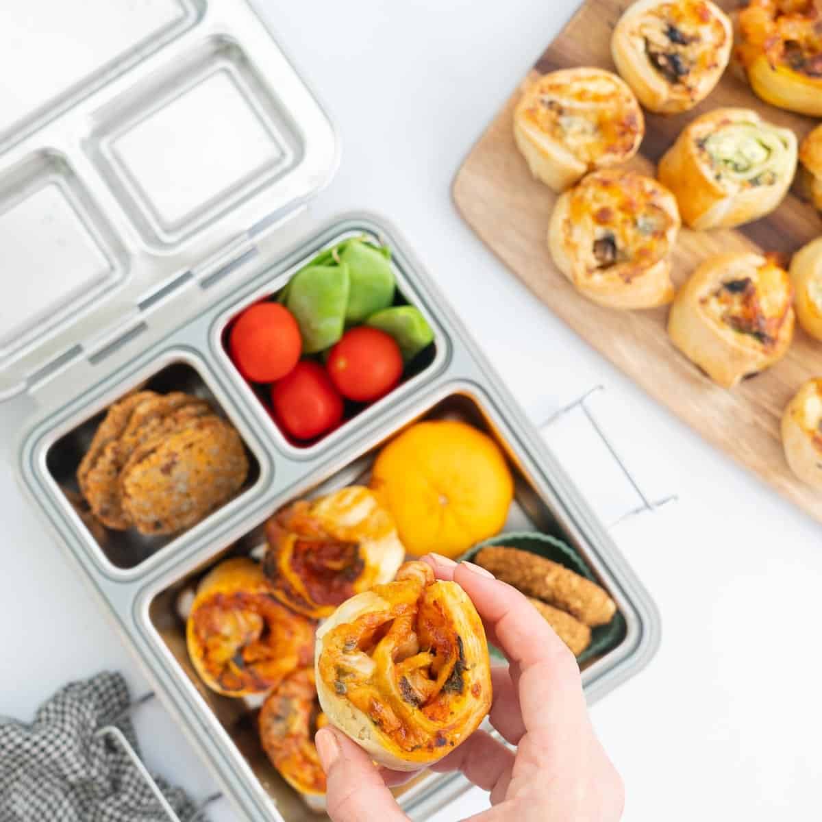 Hand holding a pizza scroll above a packed school lunch box.