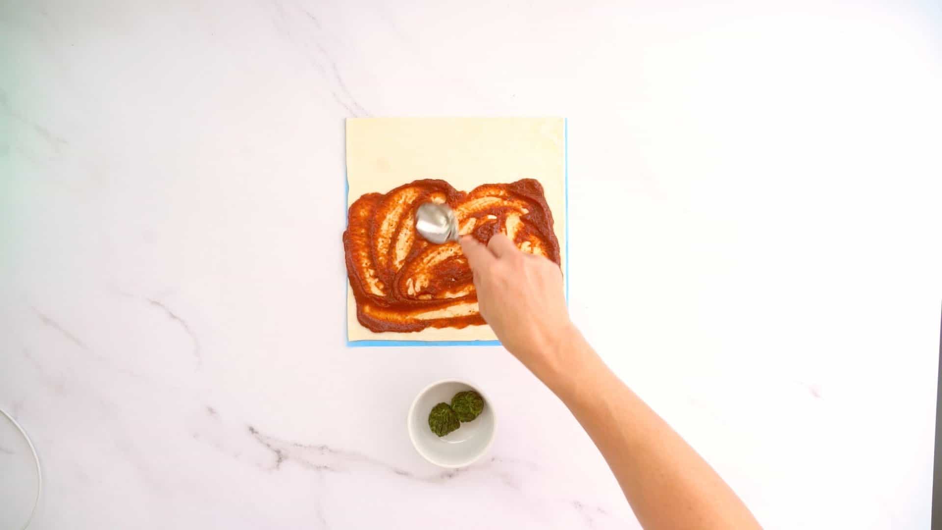 Pizza sauce being spread over a sheet of pastry.