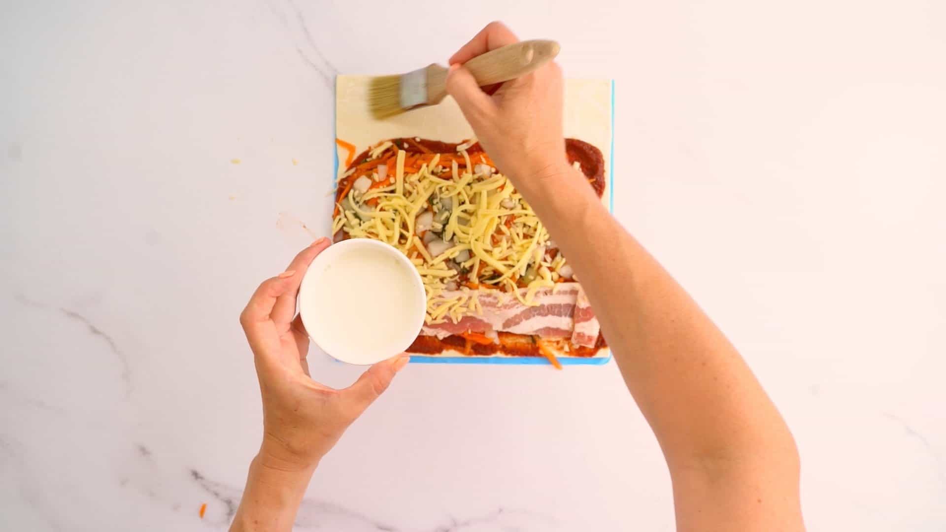 Pizza pastry topped with pizza toppings, hand brushing on milk to end of pastry with brush.