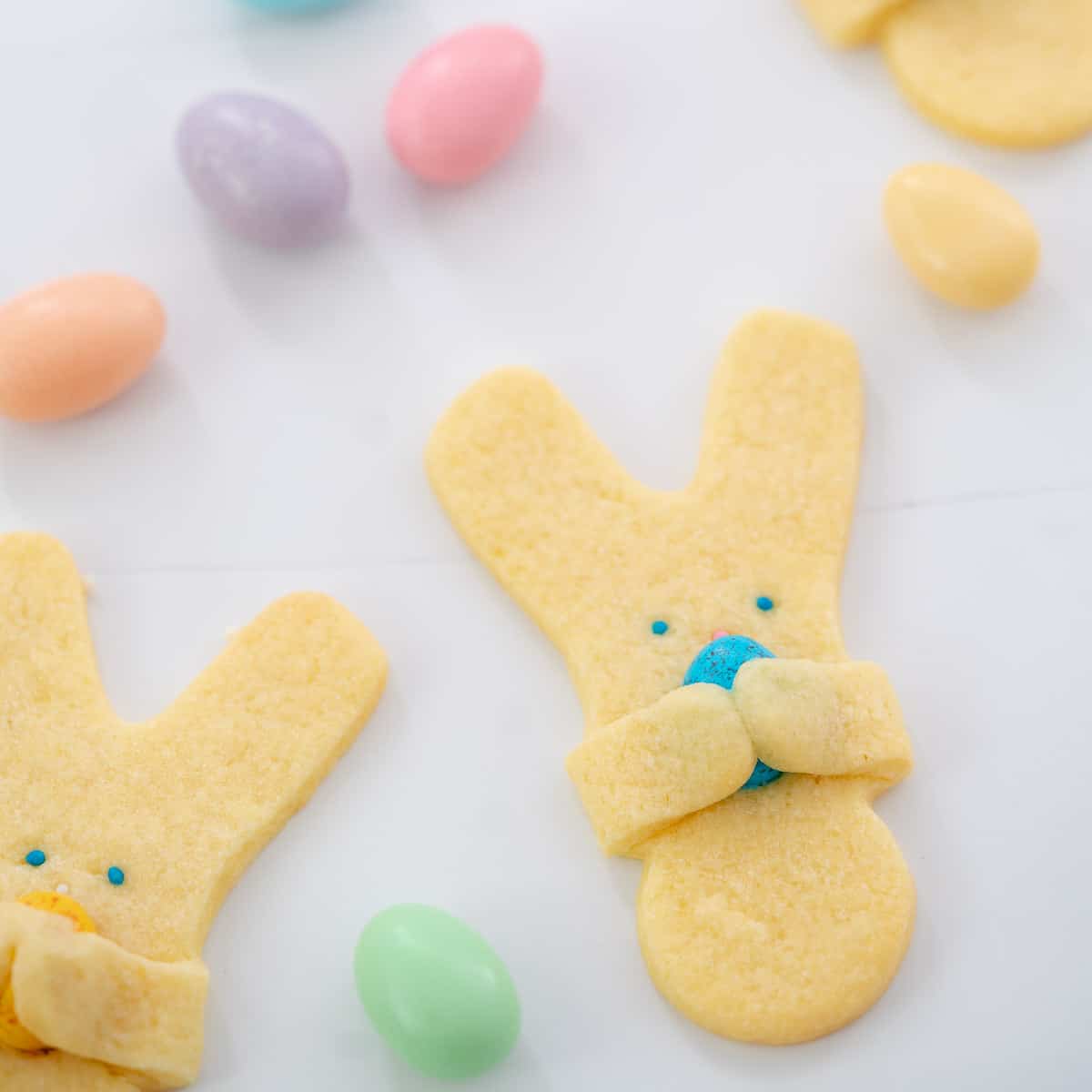 Easter bunny cookies holding mini chocolate easter eggs on a white bench top scattered with pastel coloured sugared almonds.