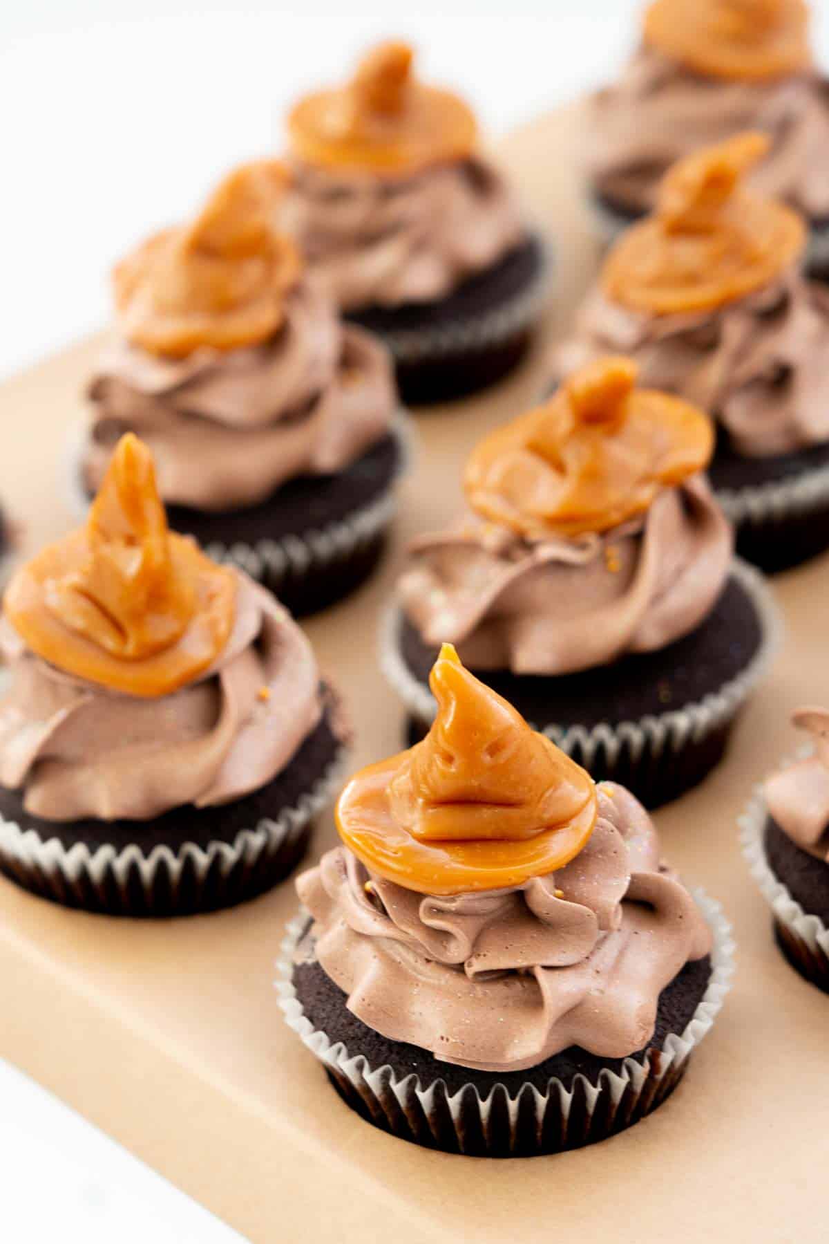 Chocolate cupcakes topped with chocolate buttercream and a caramel cupcake topper in the shape of a sorting hat from harry potter.
