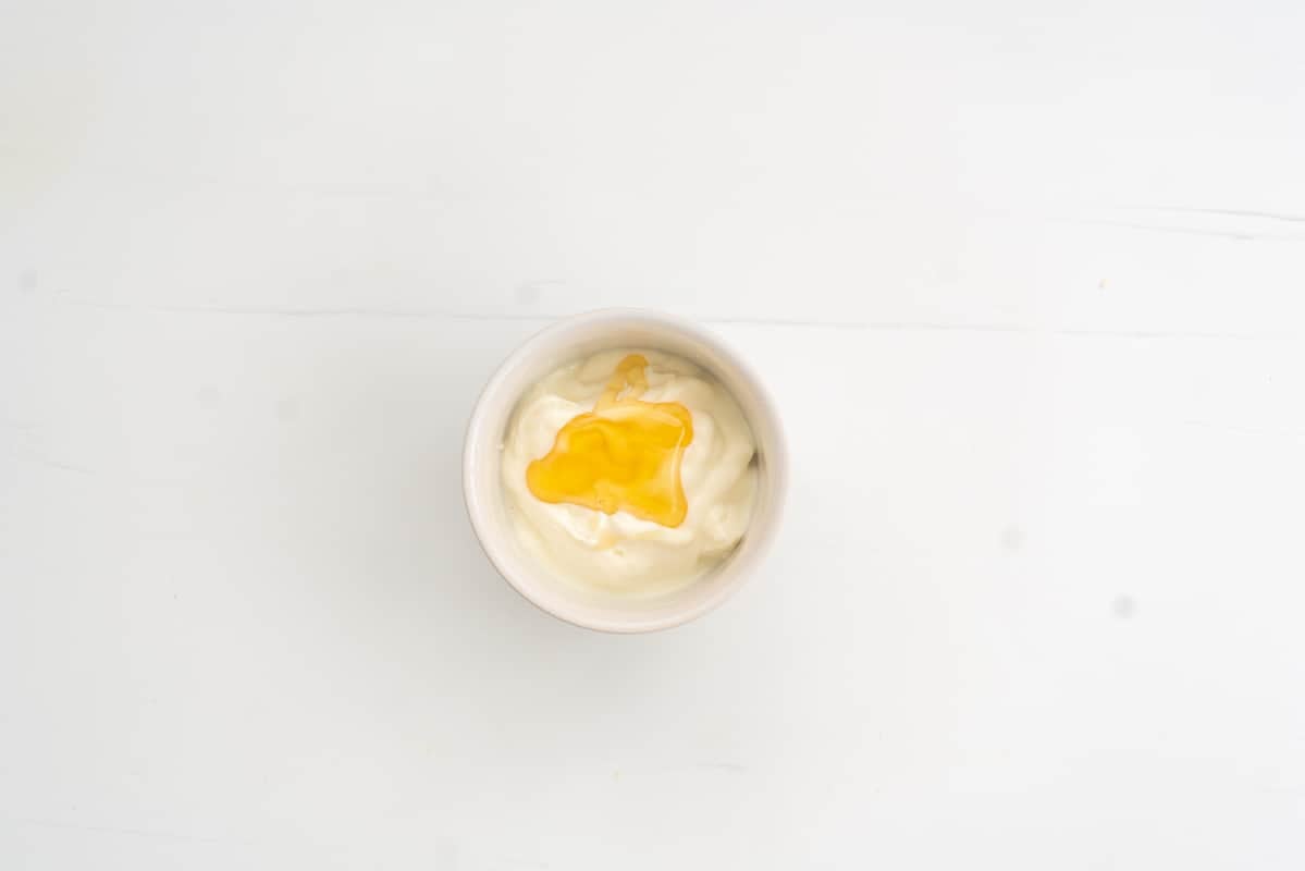 A white ceramic ramekin of greek yoghurt with a spoonful of honey on top.