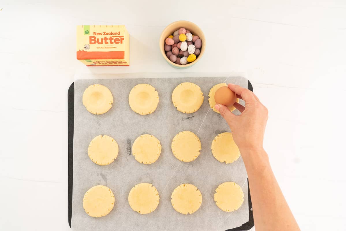An egg being pressed into cookie dough to make nest like indentations.