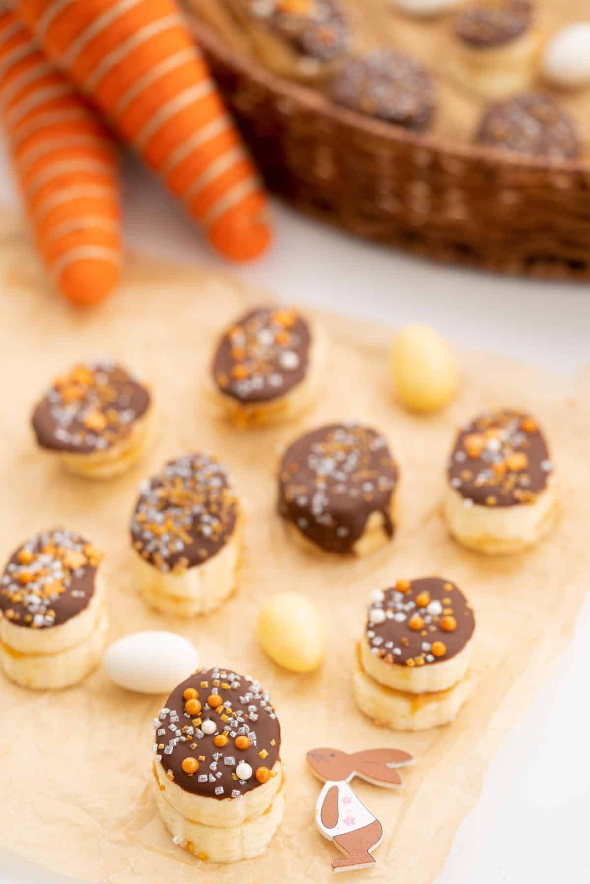 Frozen banana slices covered in chocolate and decorated with gold and silver sprinkles on brown parchment paper decorated with gold and cream easter eggs and a small easter bunny,
