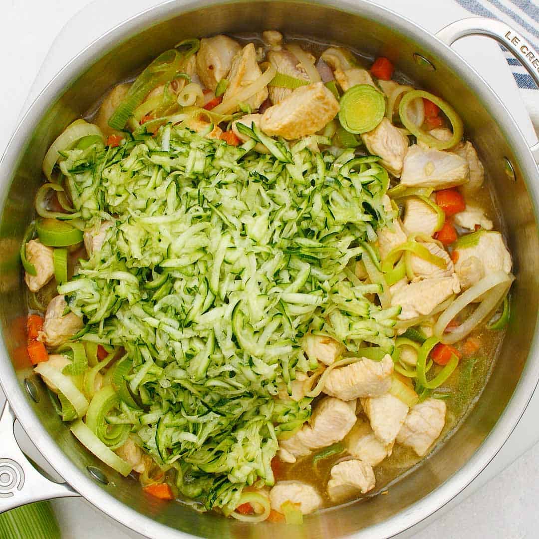 Grated zucchini added to chicken and leek cooking in a large fry pan.