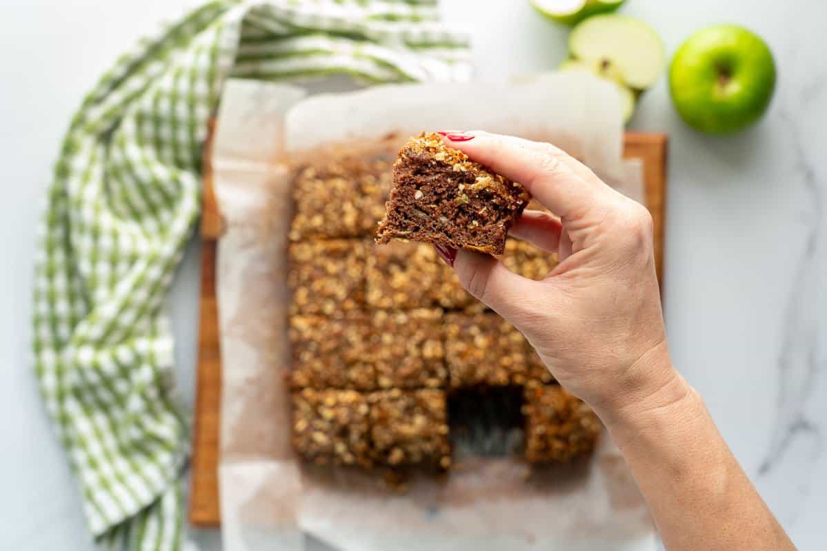 A hand holding a piece of chocolate cake above a wooden trya of more pieces of cake.