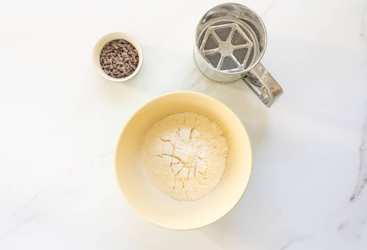 A ceramic mixing bowl with sifted in gredients on a bench top next to a sifter and small bowl of chocolate chips.