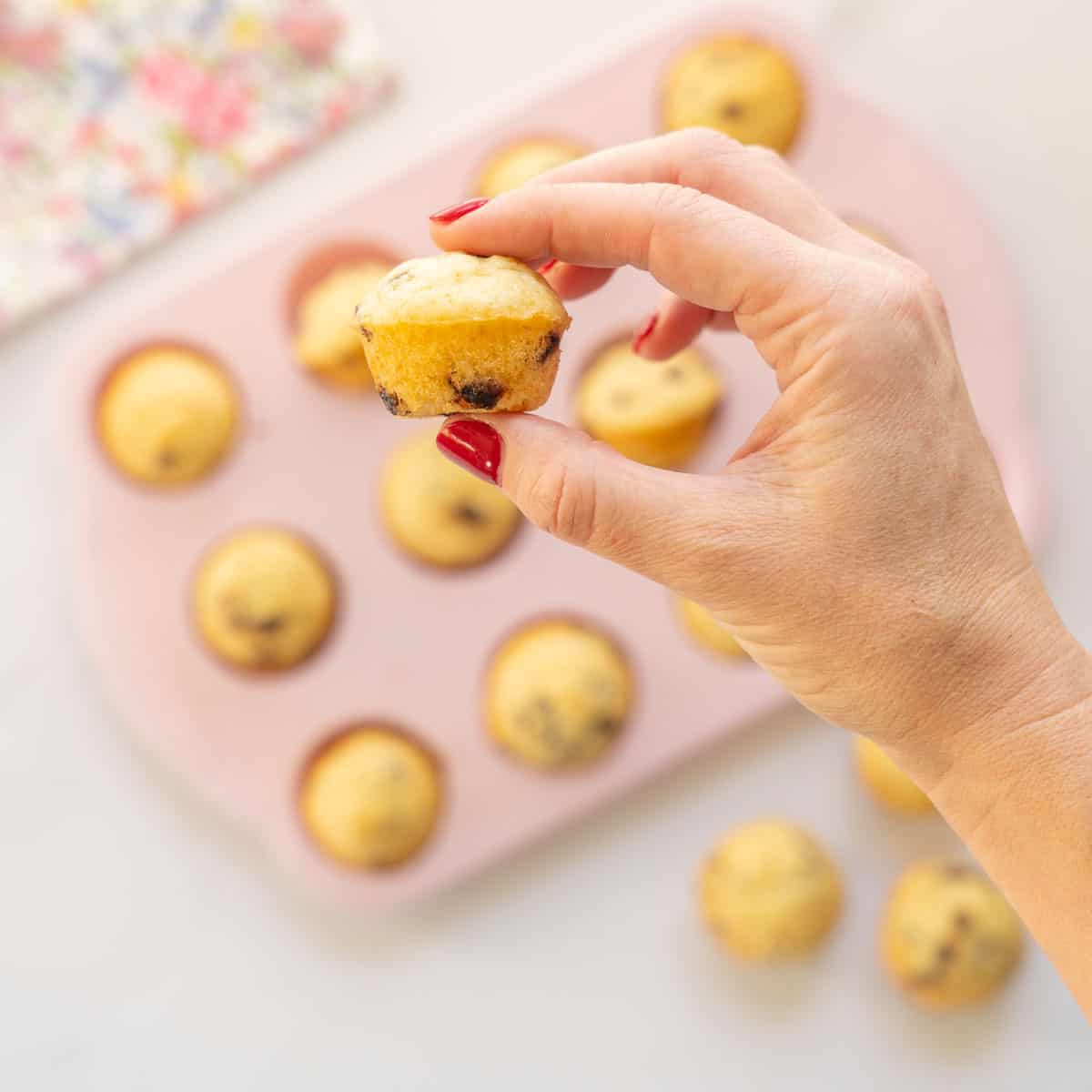 A hand holding a chocolate chip mini muffin above a muffin tray.