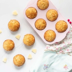 A pink muffin tray of muffins on a bench top laid out with more muffins, pineapple pieces and a floral tea towel.