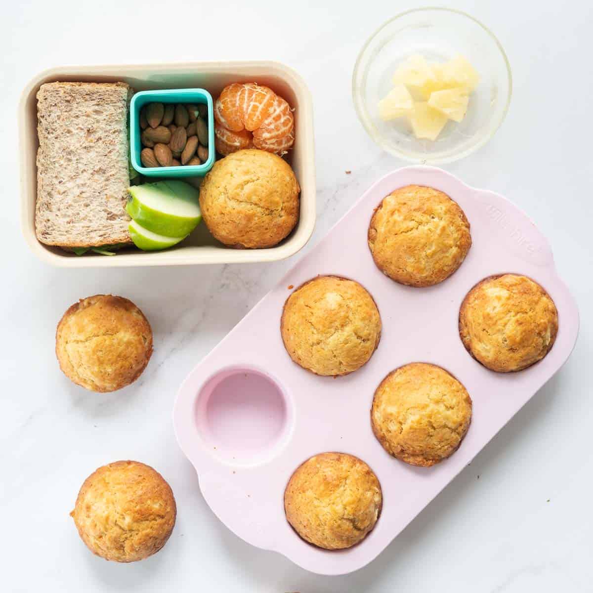 A pink muffin tray of muffins next to a packed shcool lunch box and a bowl of pineapple pieces.