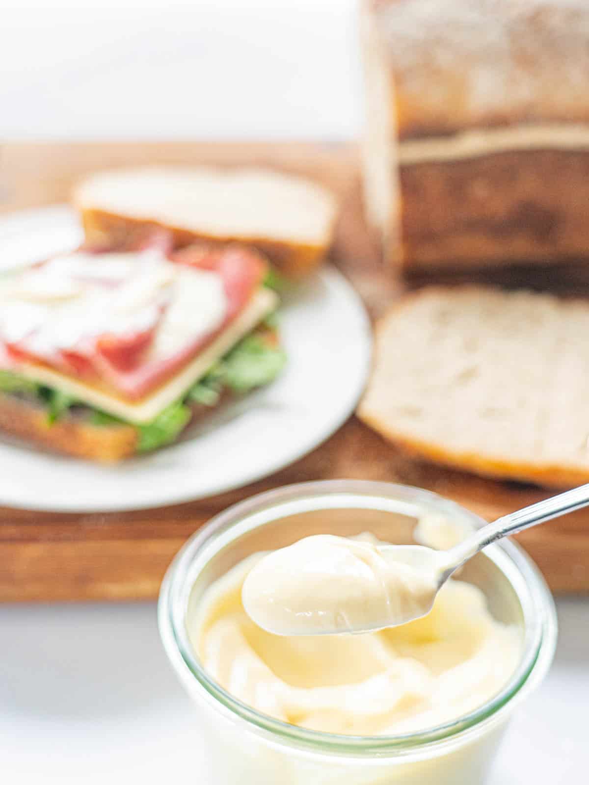 A spoonful of mayonnaise in front of a wooden board with a sandwich and loaf of bread.