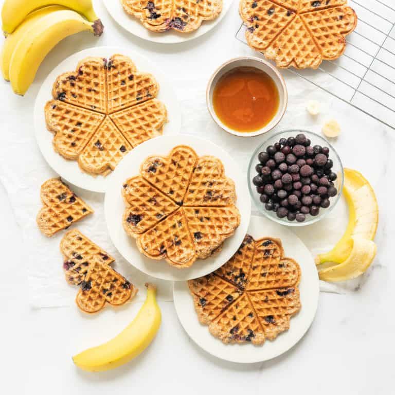 Three plates of waffles on a bench with a bowl of blueberries, maple syrup and bananas.