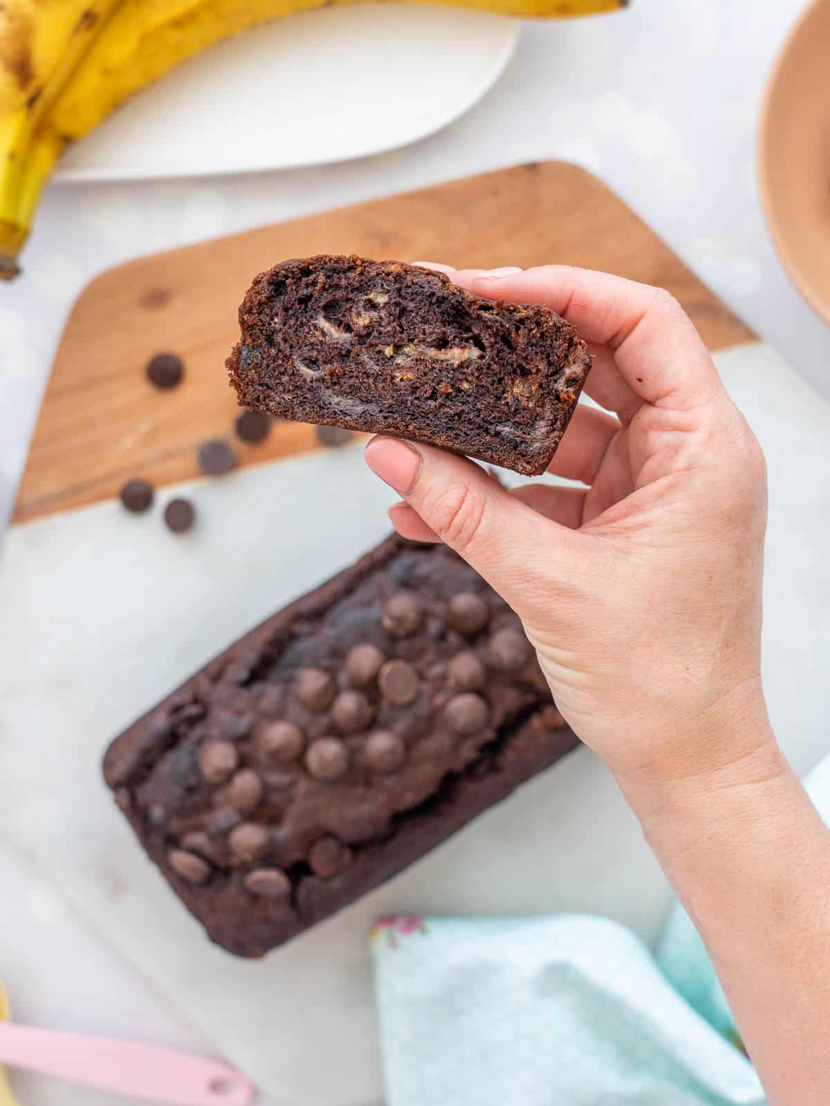A hand holding a piece of chocolate banana loaf above a marble board.