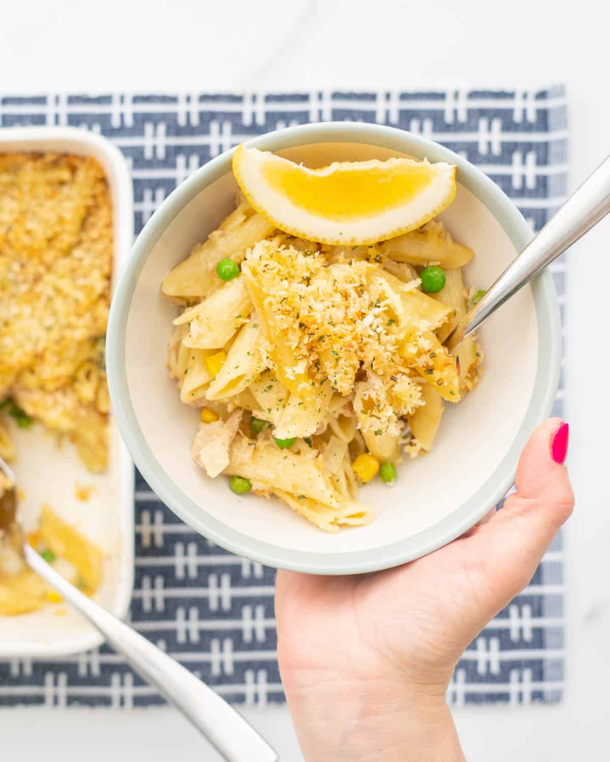 A bowl of creamy pasta topped with bread crumbs and studded with green peas held above a blue placemat.