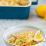 A bowl of creamy penne pasta on a grey marble bench top with a dark blue baking dish and lemons in the background.