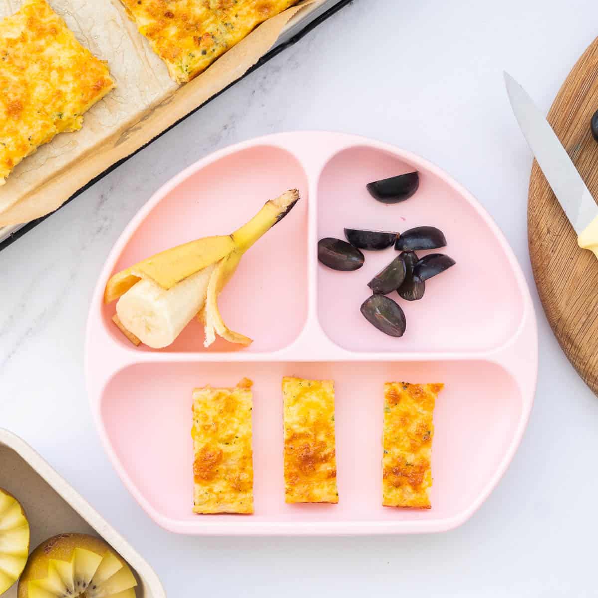 A pink silicone baby plate with strips of vegetable slice, banana and quartered grapes.