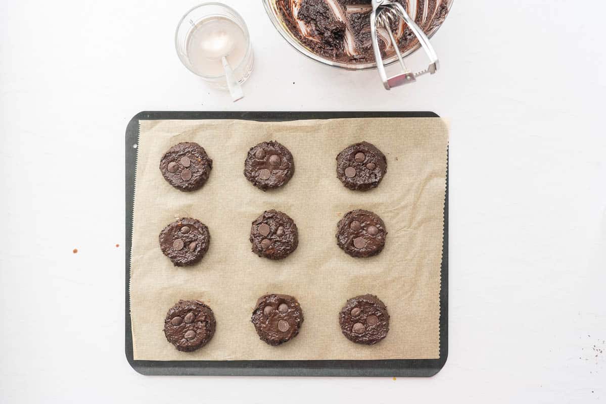 Nine chocolate cookies studded with chocolate drops on a baking paper lined tray ready to go into the oven.