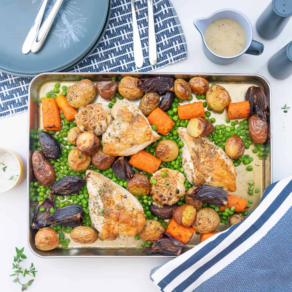 A roast chicken tray bake on a table set with blue placemats, plates, cutlery and a gravy boat.