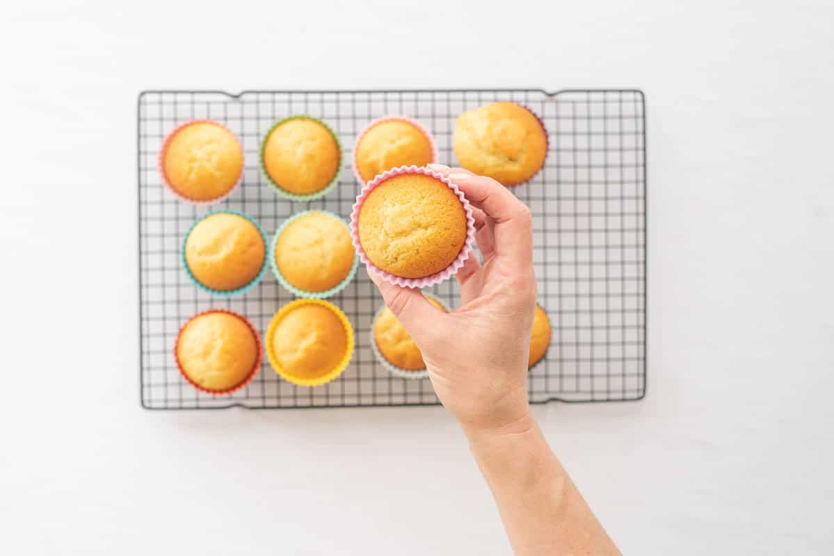 A hand holding a golden cupcake in a pink case above a cooling rack of more cupcakes.