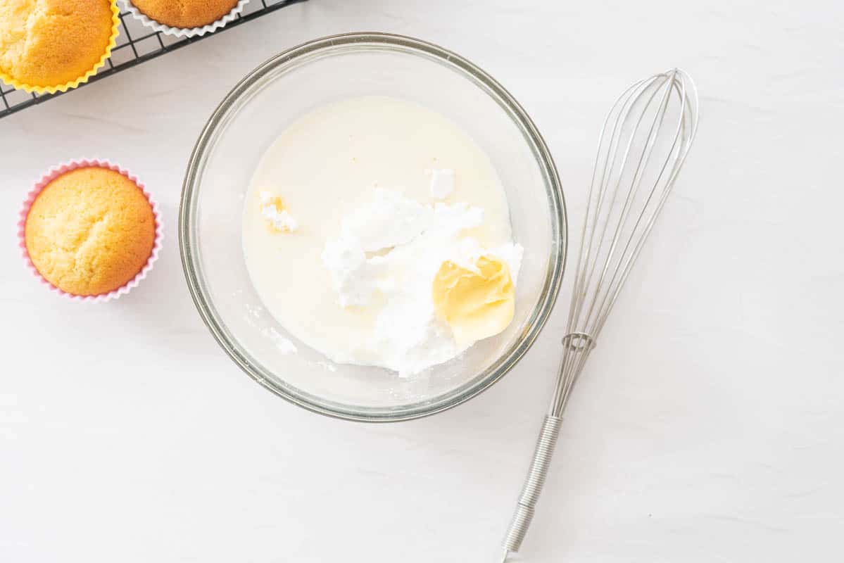 A glass mixing bowl with vanilla extract, butter, milk and icing sugar next to a whisk.