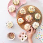 A child's hand reaching for cupcakes decorated with white frosting and sprinkles on a wooden chopping board.