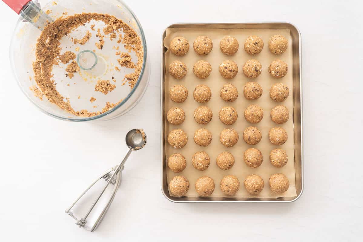 Banana energy bites on a lined baking tray next to a cookie scoop and empty food processor bowl.