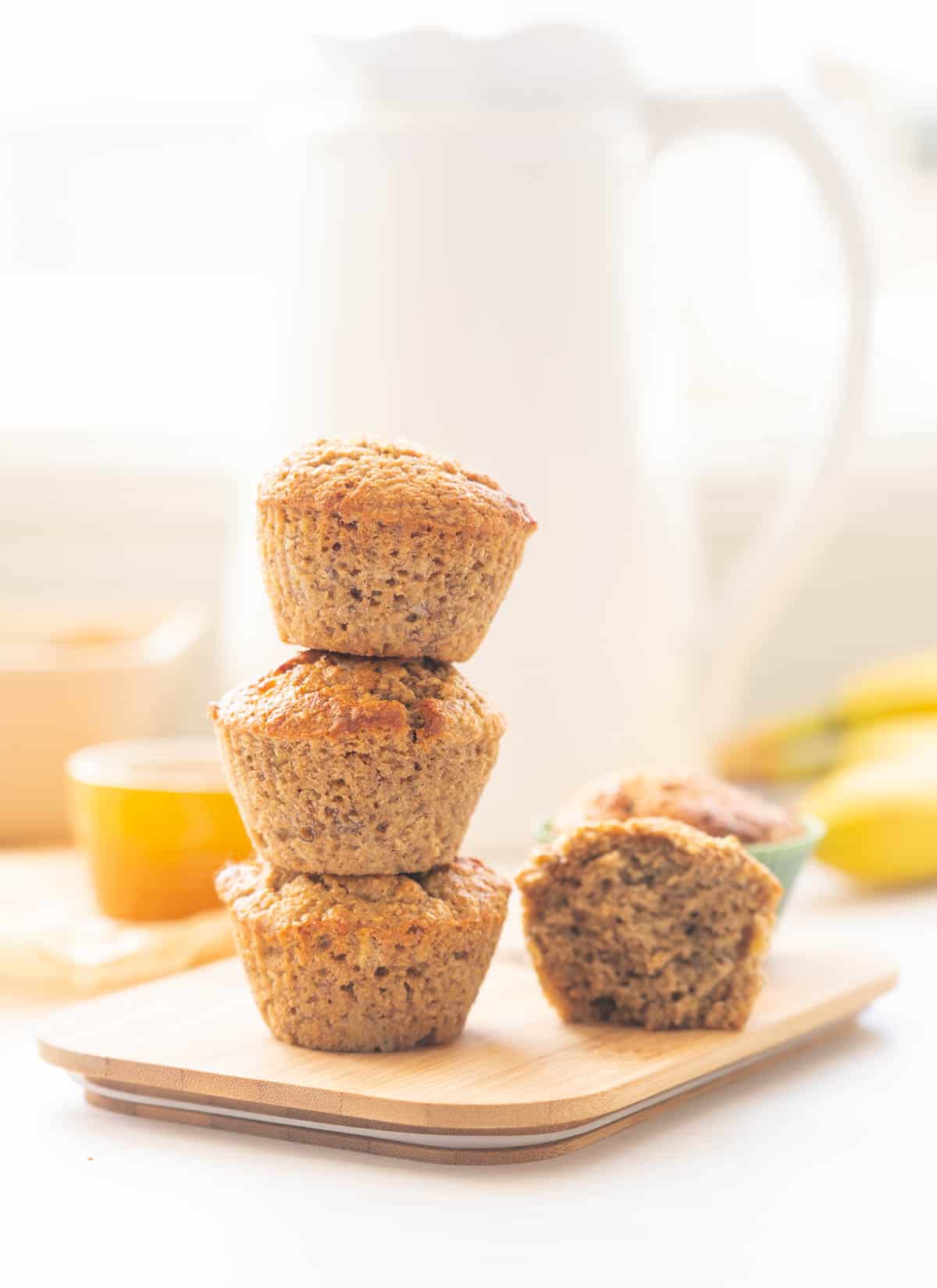 A tower of three bran muffins on a wooden board sitting in front of a white jug.
