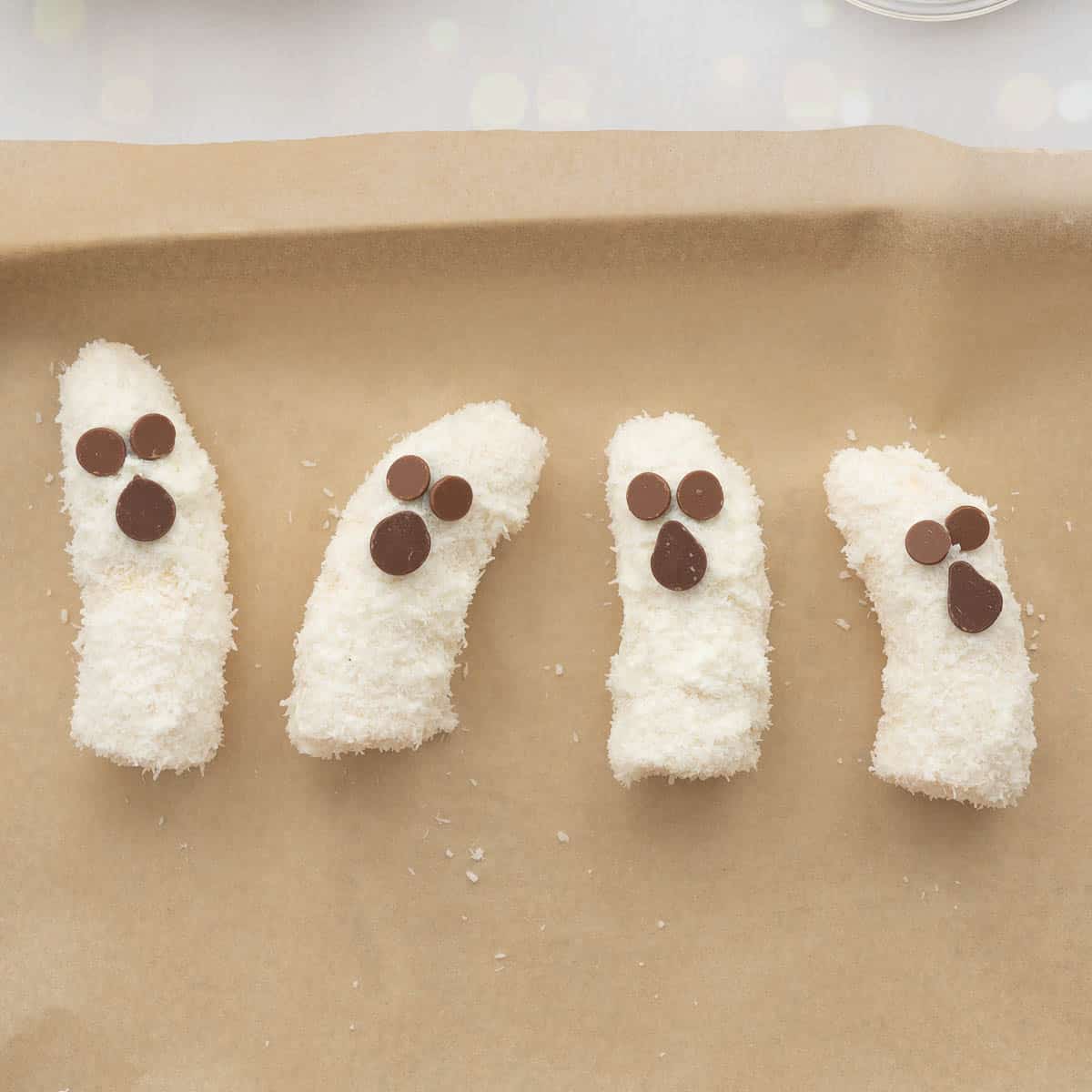 4 banana halves decorated with desiccated coconut and chocolate drops to look like chocolate drops lying on a lined tray.