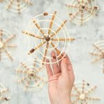 A spider web made from pretzel sticks and white chocolate, and decorated with a milk chocolate spider in the centre being held up.