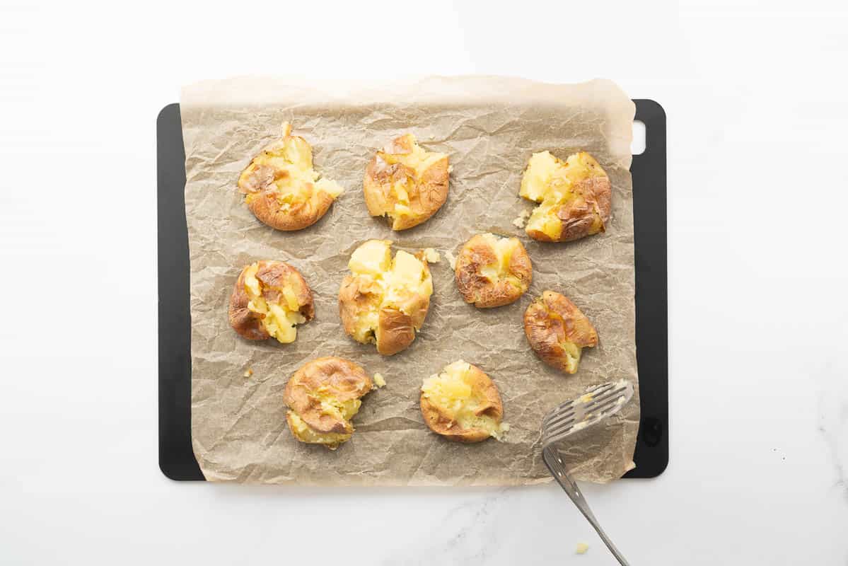 Flattened potatoes on a chopping board with a potato masher lying to the side.