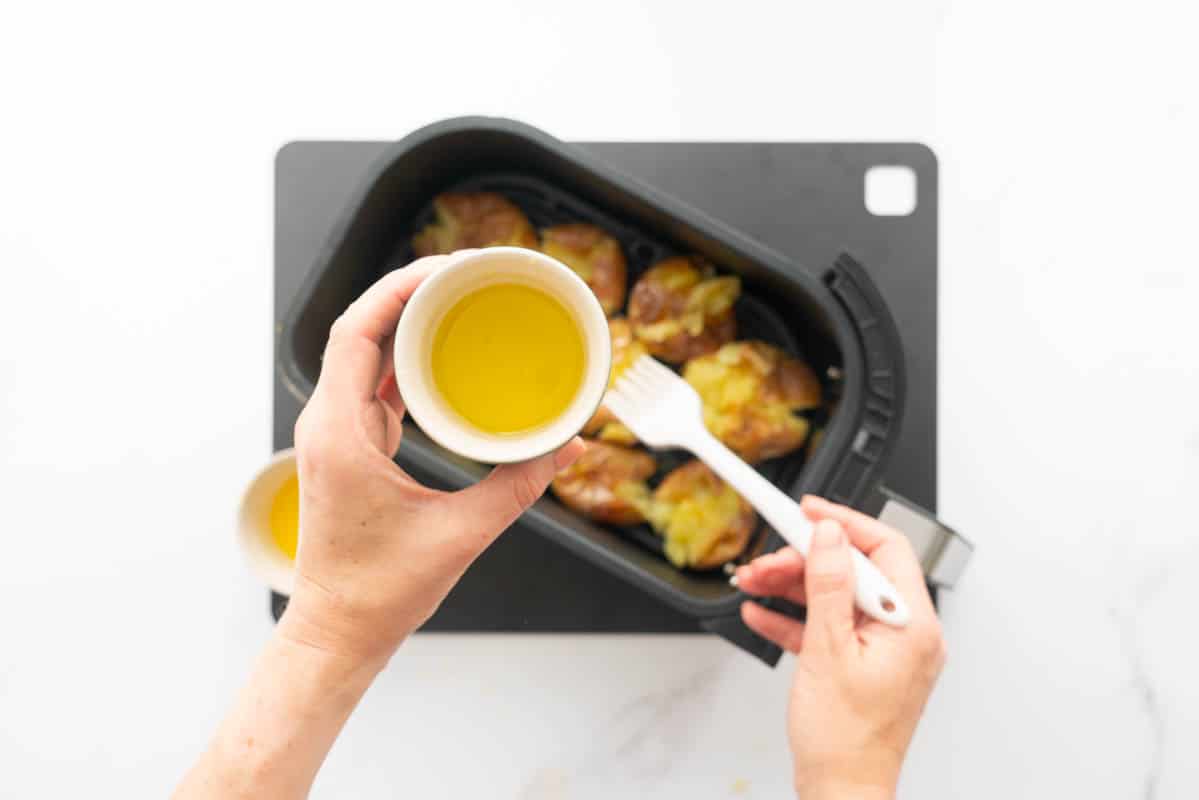 Crushed potatoes in an air fryer basket being brushed with olive oil.