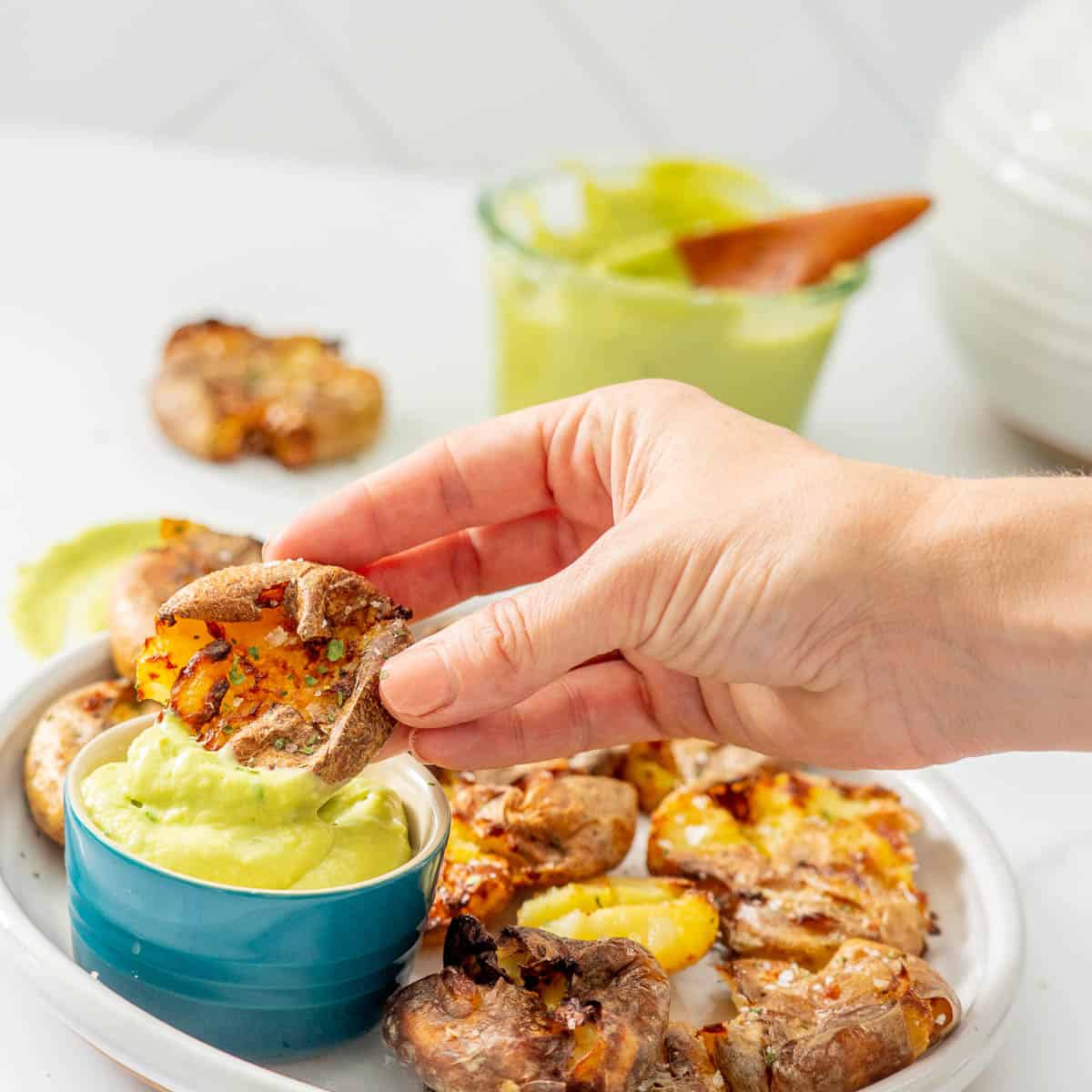 A crispy smashed potato being dipped into a small blue bowl of avocado aioli.