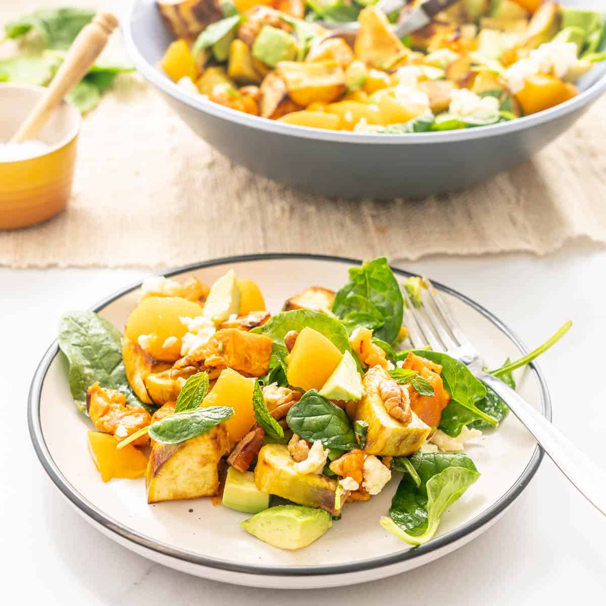 Salad on a black rimmed cream ceramic plate, sweet potato, avocado, spinach, peaches, peans and feta visible.