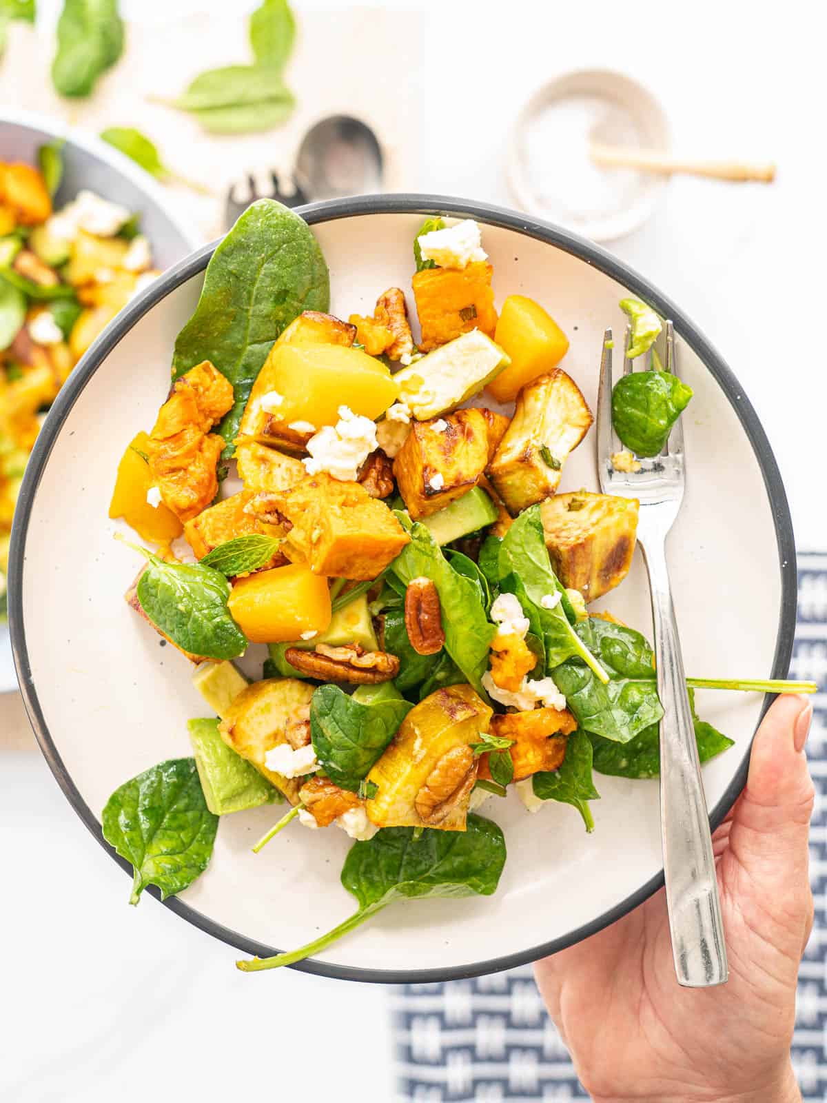 A plate of salad containing sweet potato, peaches, pecans, feta, avocado and spinach leaves being held by a hand with a fork resting on the side of the plate
