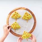 Two children's hands reaching out to pick up 4 triangular pieces of pita bread decorated to look like Christmas trees.