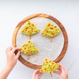 Two children's hands reaching out to pick up 4 triangular pieces of pita bread decorated to look like Christmas trees.