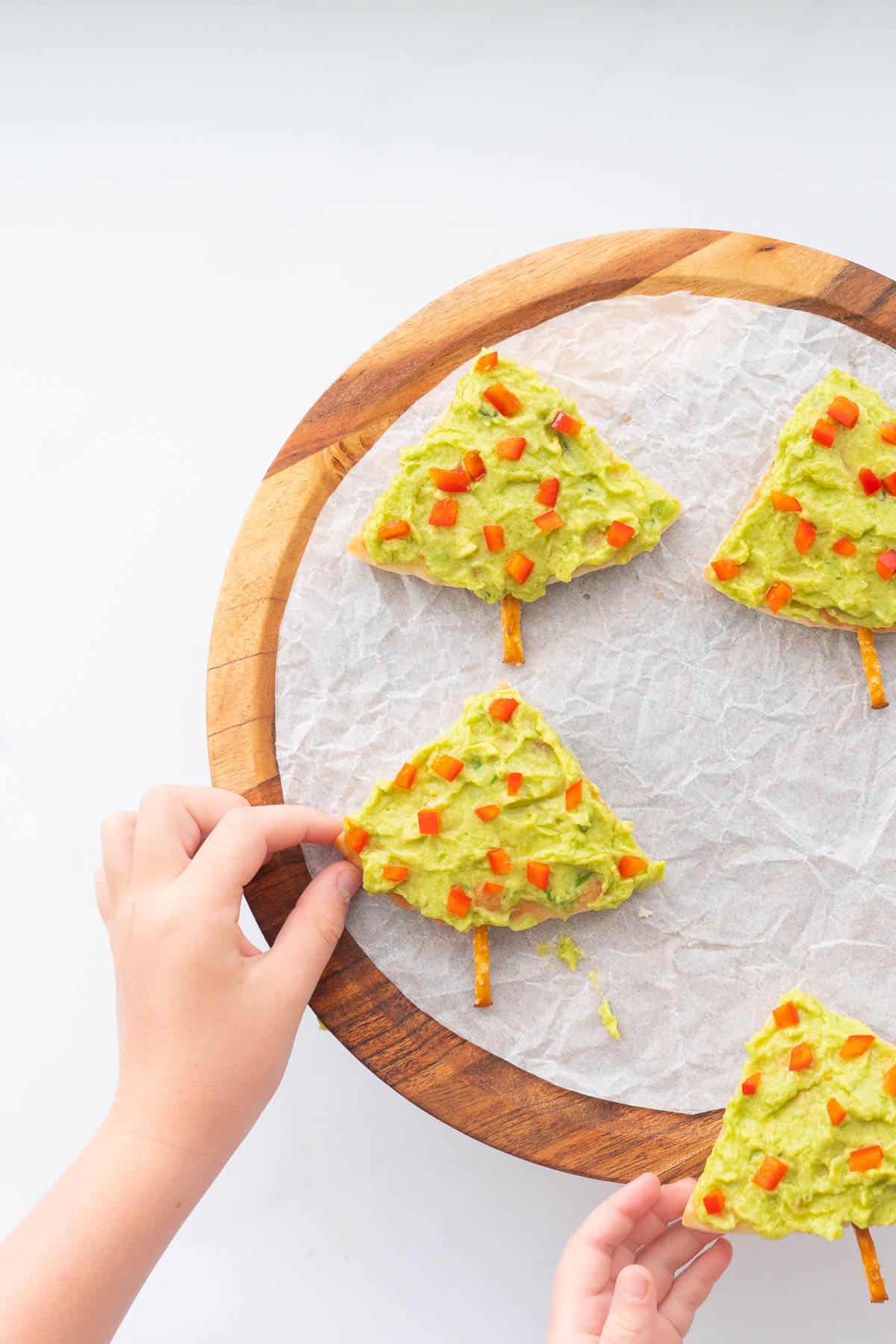 Two children's hands reaching out to pick up 4 triangular pieces of pita bread decorated to look like Christmas trees.